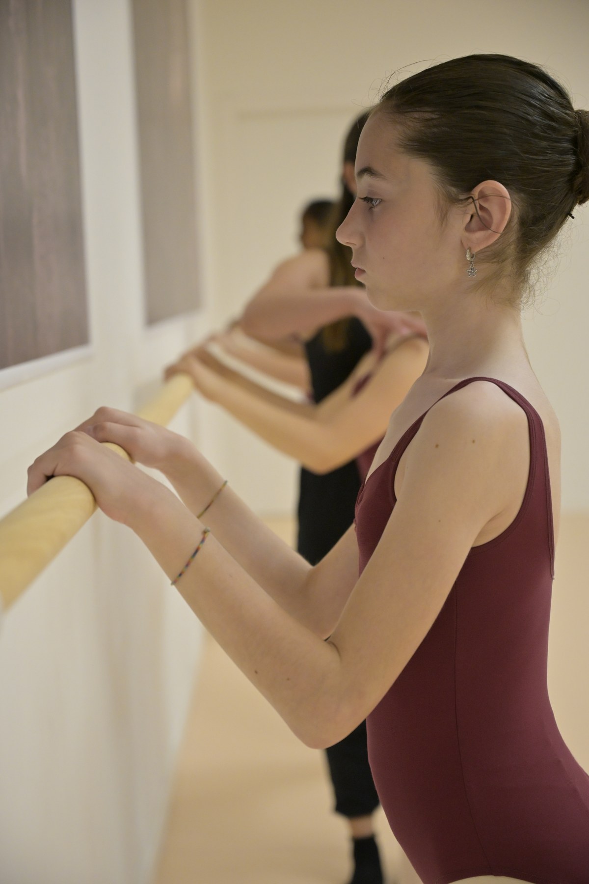 Ragazza con chignon e body bordeaux alla sbarra di legno chiaro, con altre allieve in fila nella sala