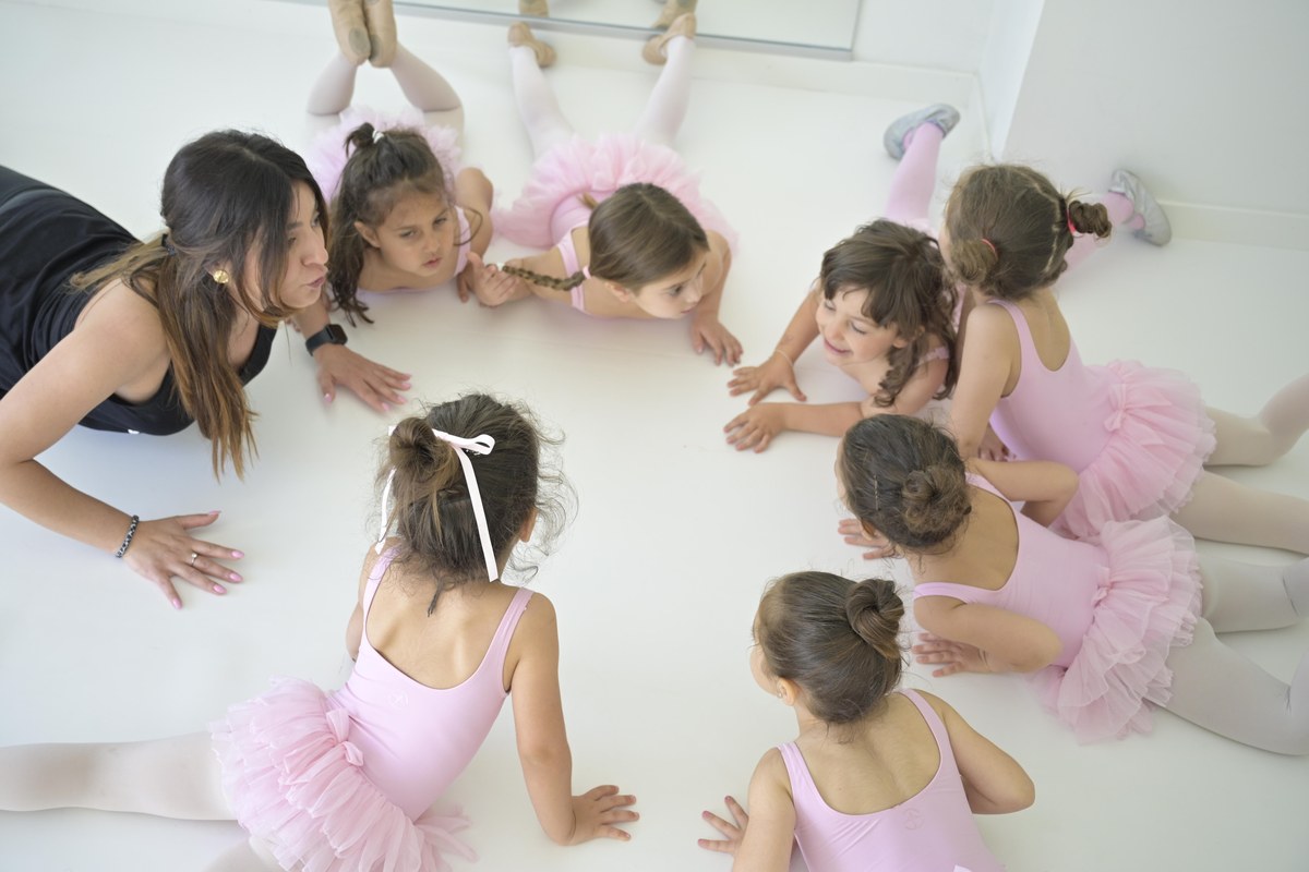 Insegnante e sette bambine in body e tutù rosa sdraiate in cerchio su un pavimento bianco da Taraxè Ballet Academy
